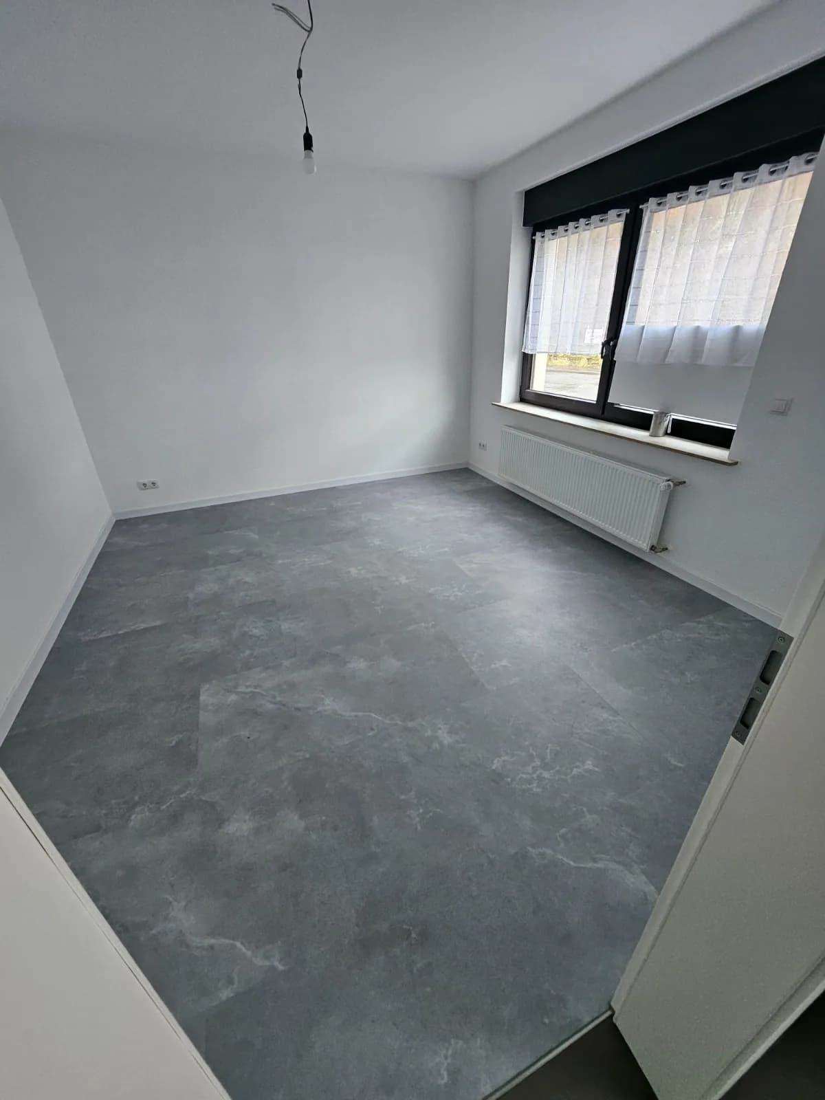 Zimmer mit grauem Bodenbelag und gestrichenen Wänden am großen Fenster